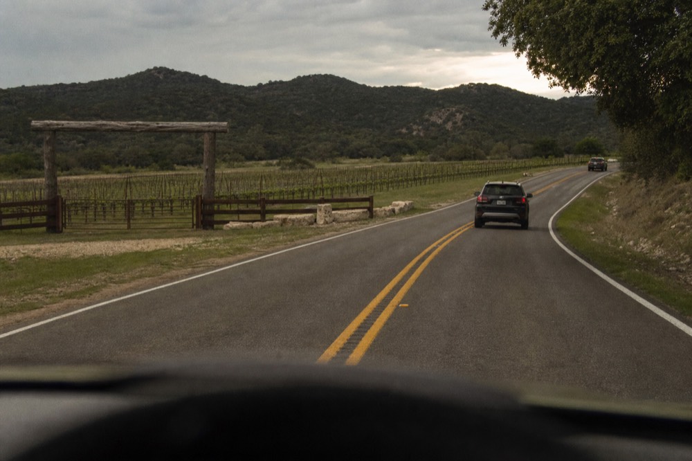 A two-lane highway curving between low hills with vine rows on both shoulders and a wooden gate marking a property entrance