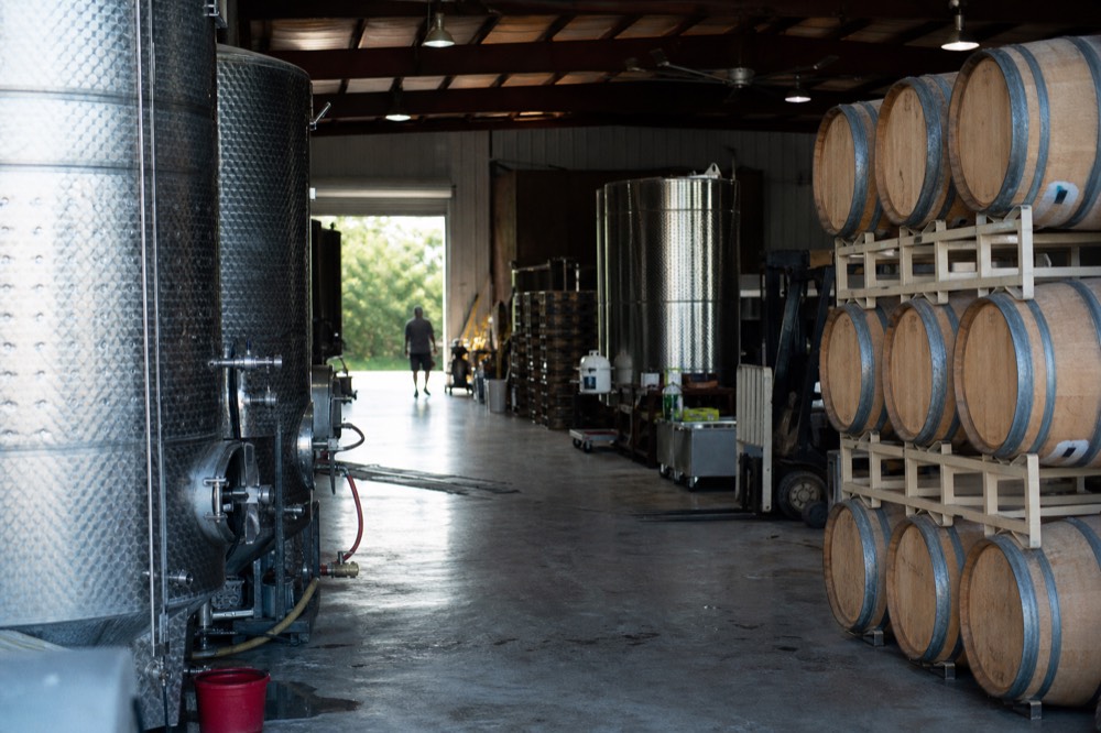 Stainless steel fermentation tanks and stacked oak barrels inside a small working winery with concrete floors and roll-up doors