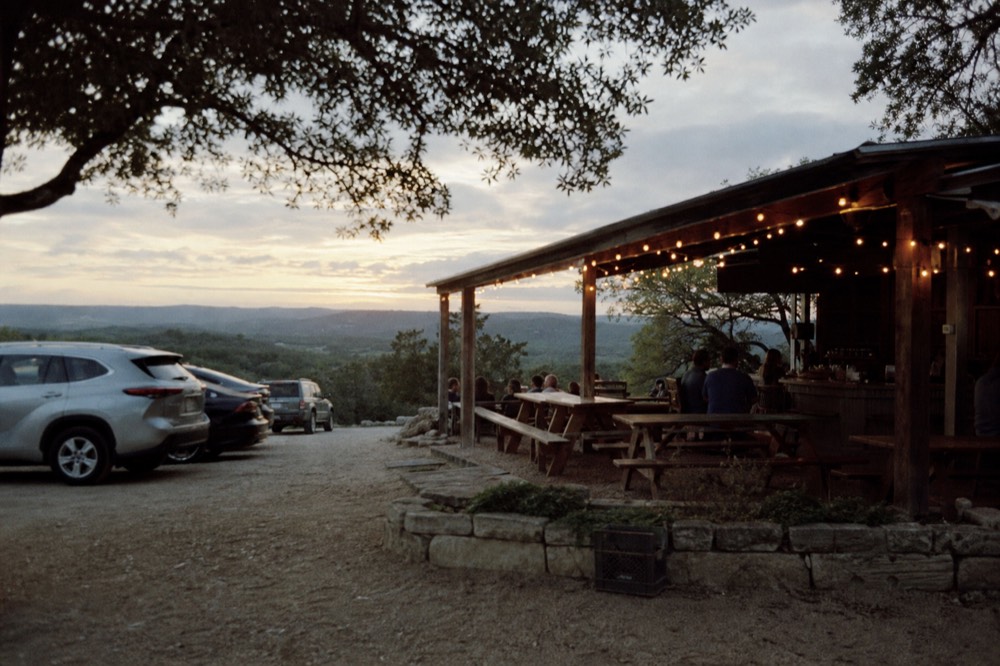 A rustic open-sided bar under a corrugated roof with wooden picnic tables, string lights and dappled shade from overhead oaks