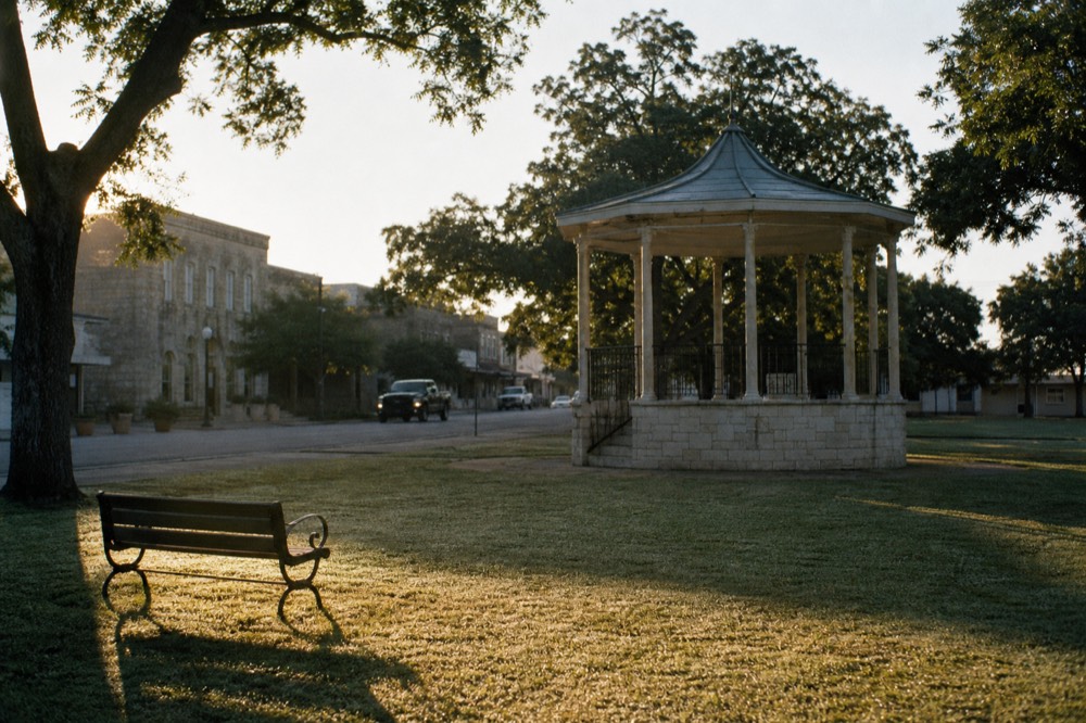 An empty town square at sunrise with an eight-sided stone landmark, dew on the grass and a single bench in the foreground