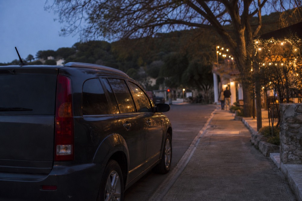 A car parked on a quiet small-town side street at dusk with warm light spilling from a restaurant doorway across the pavement