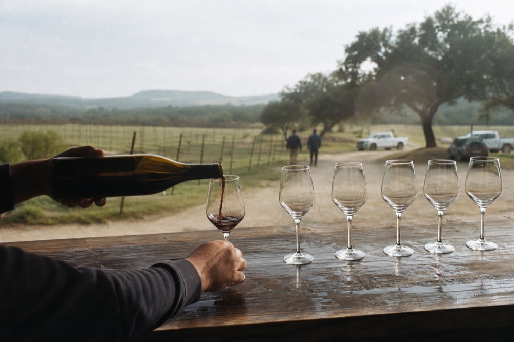 A pourer's hands tipping a bottle into a small tasting glass across a wooden bar, other glasses waiting in a row