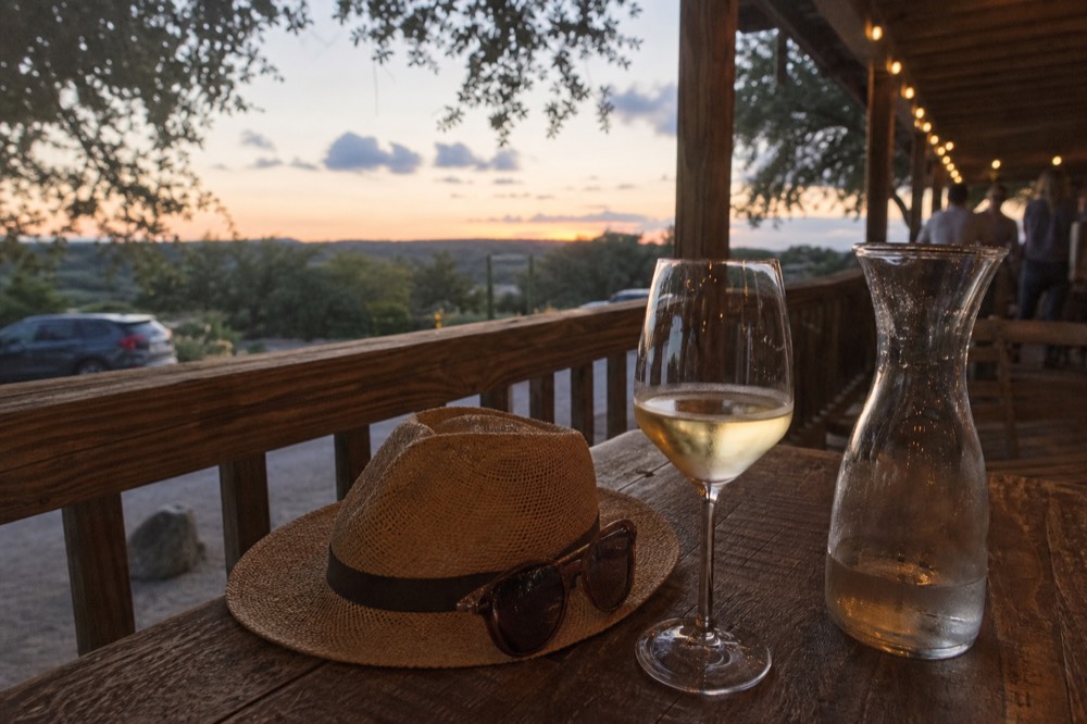 A wide-brim straw hat and sunglasses resting on a wooden patio table beside a glass of white wine and a water carafe