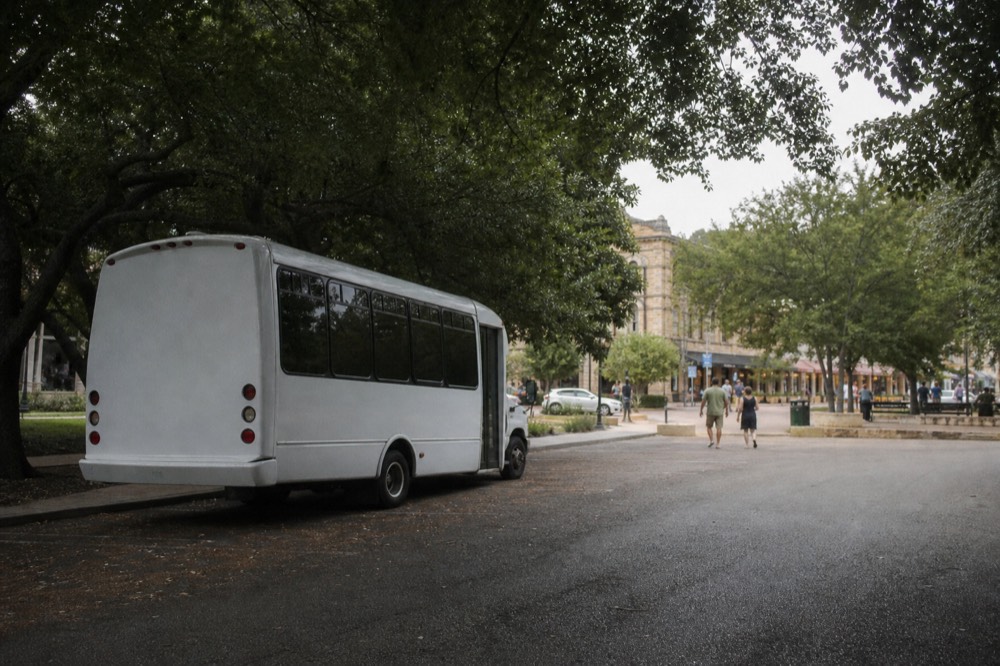 Small tour bus parked in a shaded lot near a historic Texas town square with couples walking toward the shops