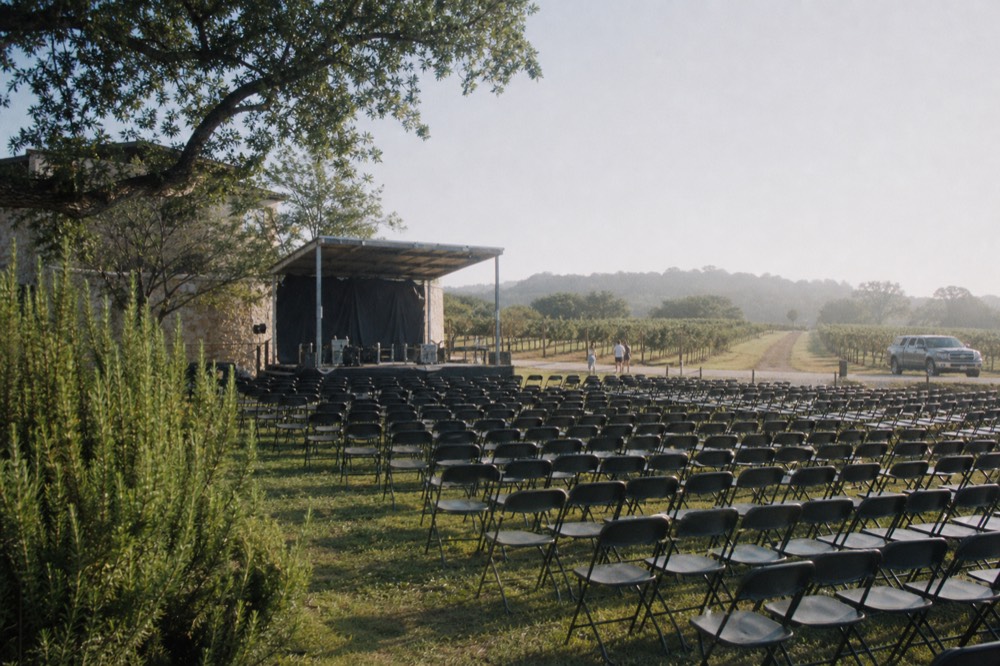 Rows of empty folding chairs and a small covered stage set up on grass beside a stone building, morning light before an event opens
