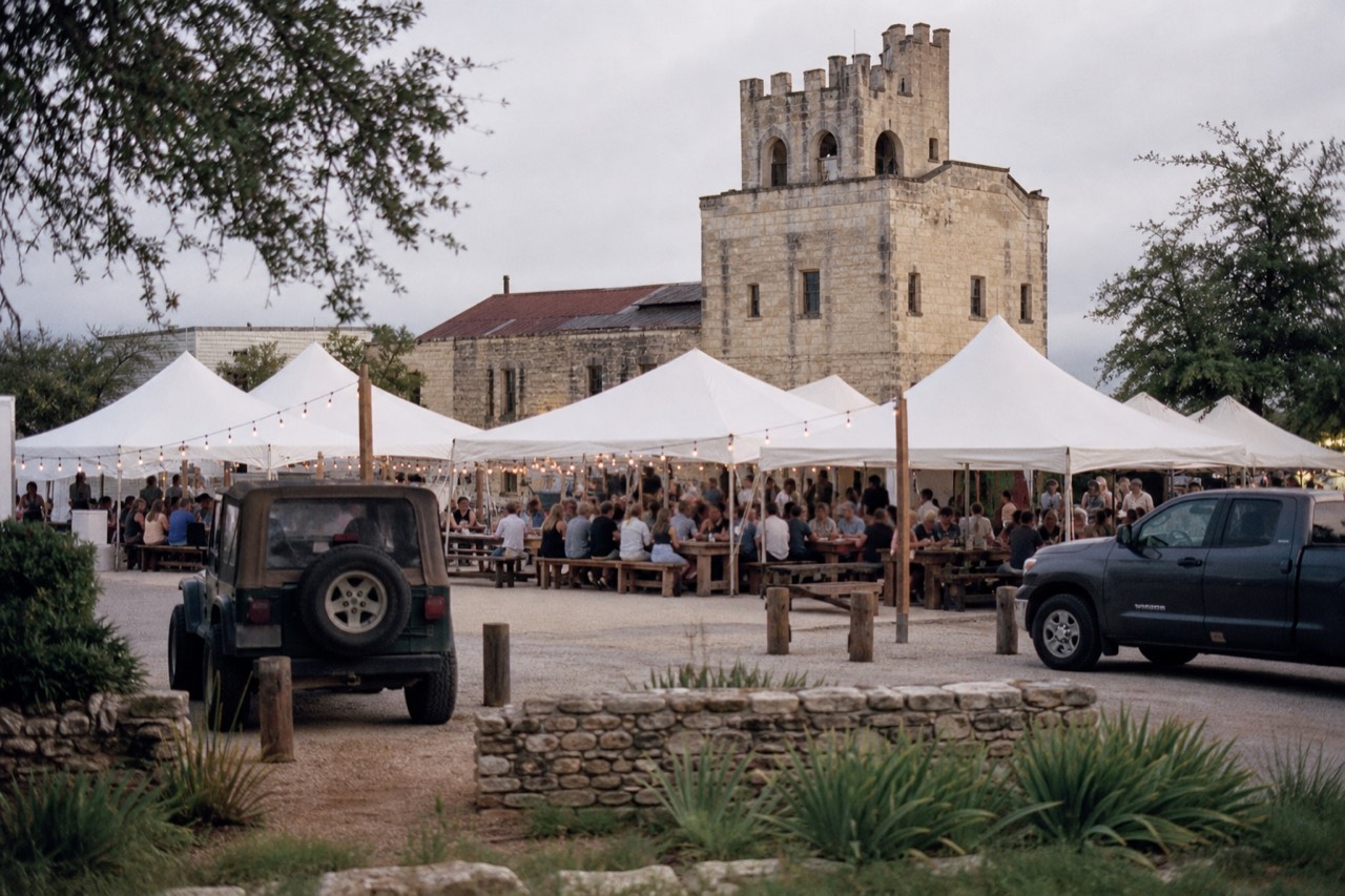 An open town square filled with white event tents and long tables at dusk, string lights overhead, a tall stone tower building behind
