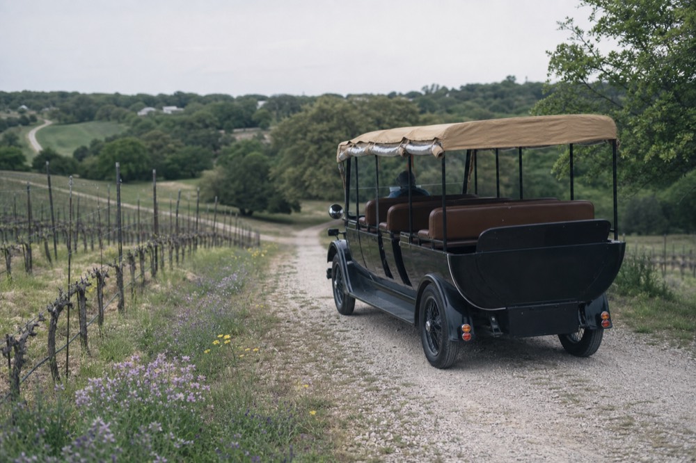 An open-air vintage-style touring vehicle with bench seats parked on a gravel drive beside vineyard rows in the Texas Hill Country