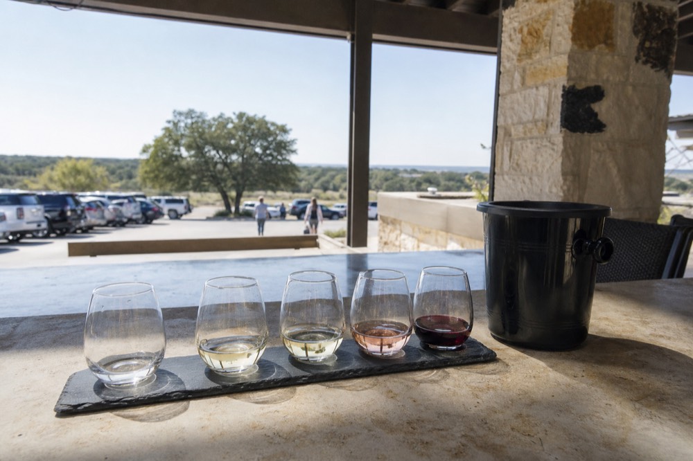 A tasting bar counter with five small pour glasses lined up on a slate flight board and a spit bucket to one side