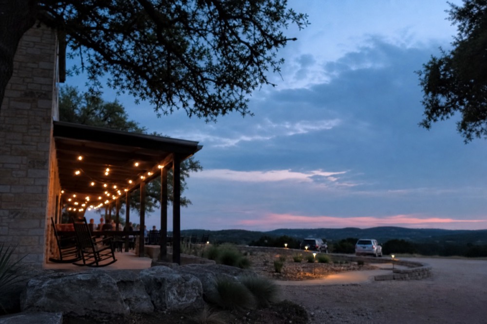 A limestone Hill Country guesthouse with a wide covered porch, rocking chairs and string lights at dusk