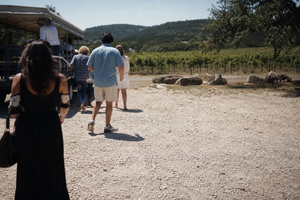 A small group stepping down from an open-sided vintage tour vehicle onto a gravel lot beside a vineyard, late morning shadows