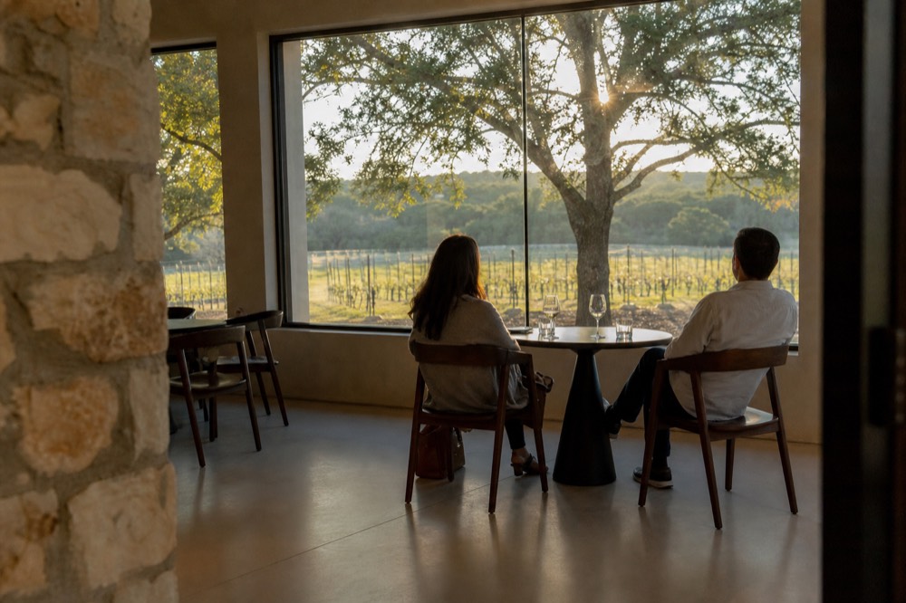 Two people seated at a small round table in a near-empty tasting room, glasses half full, wide window showing vine rows outside