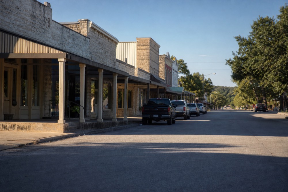 Limestone storefronts with deep porches along a wide Texas main street, striped awnings and parked pickup trucks in soft late-afternoon light