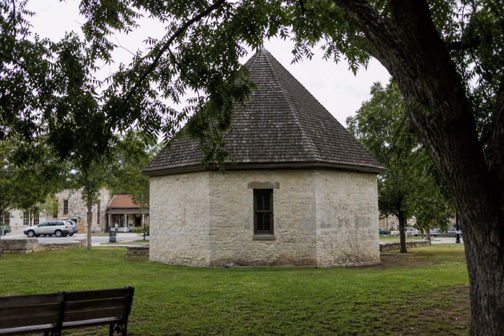 An eight-sided stone building with a steep shingled roof standing on a green town square surrounded by oak trees and benches