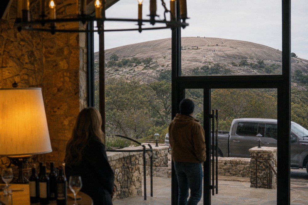 A vast pink granite dome rising out of scrub oak and grassland, with tiny figures walking up the bare rock face
