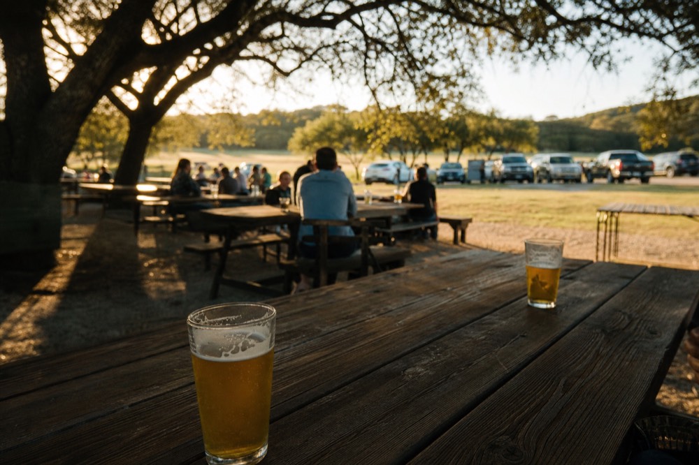 A shaded biergarten with long communal wooden tables, gravel underfoot and glasses of pale beer catching the late light