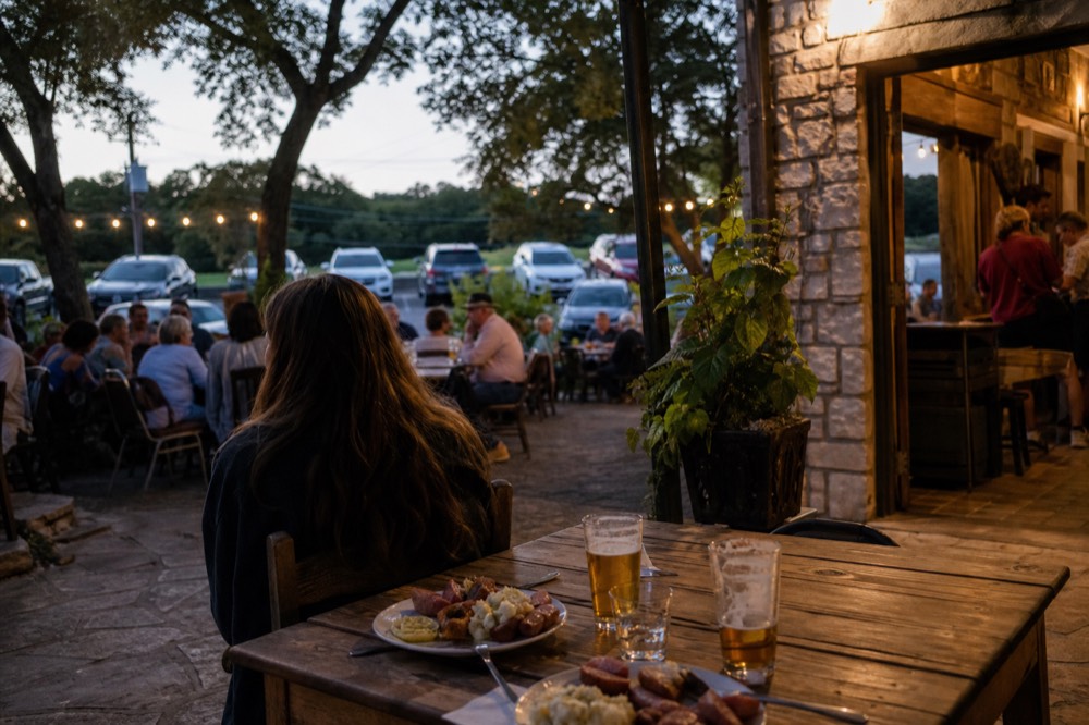 A busy restaurant terrace at dusk with wooden tables, half-full beer glasses and plates of sausage and potato, warm light spilling from a doorway