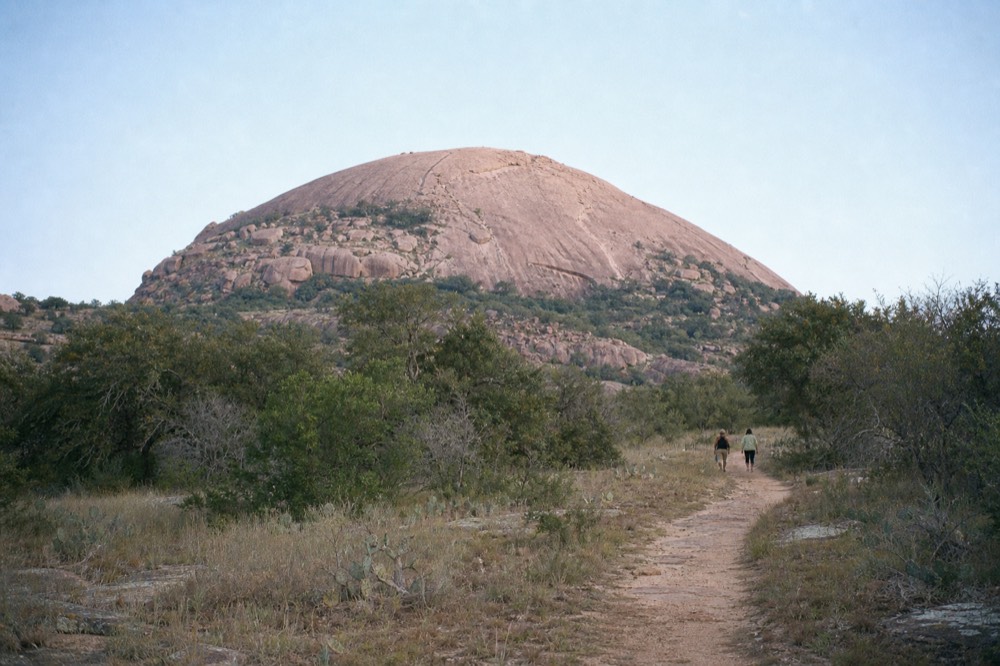 A vast pink granite dome rising above scrubby oak and grass under clear morning sky, a faint worn trail line curving up the slope