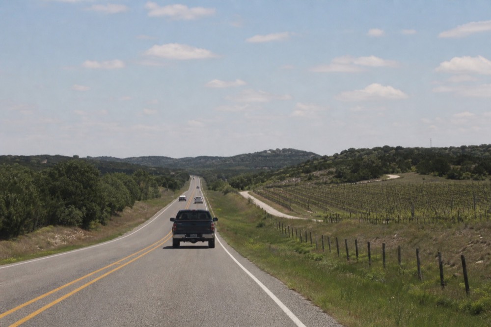 A straight two-lane highway running between low hills and vineyard rows, wooden fence posts along the shoulder, midday sun