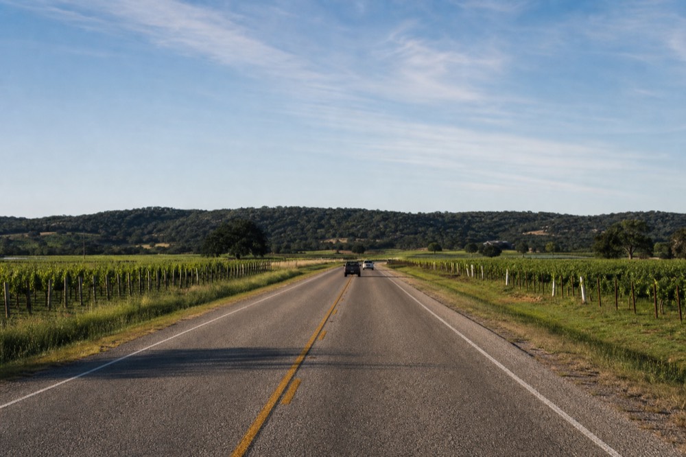 A long straight stretch of two-lane highway with vineyard rows on both sides and a distant line of hills, mid-morning light