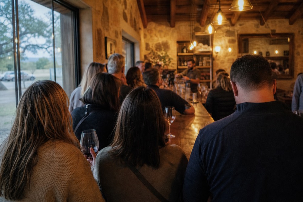 Crowded winery tasting bar on a weekend afternoon seen from behind waiting guests, staff pouring at the far end
