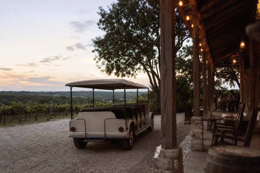 Open-air vintage shuttle parked on gravel beside a Hill Country vineyard row in late-afternoon light