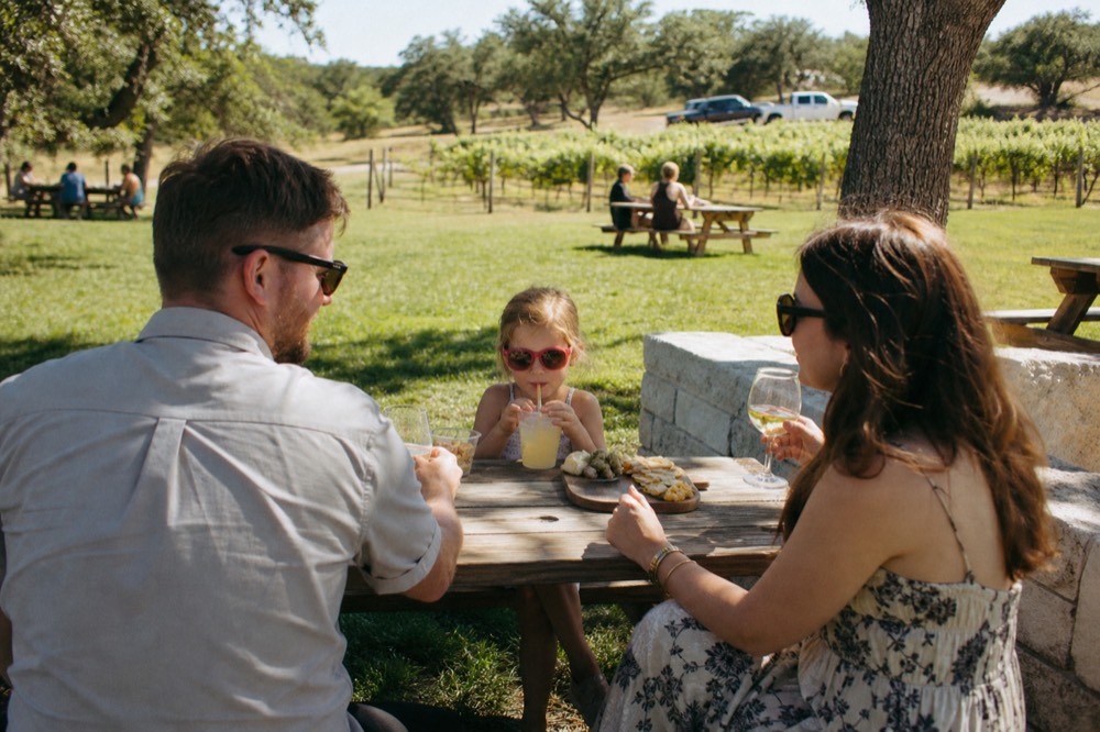 Family group seated at an outdoor picnic table on a winery lawn, adults with glasses, a child with lemonade, vines behind