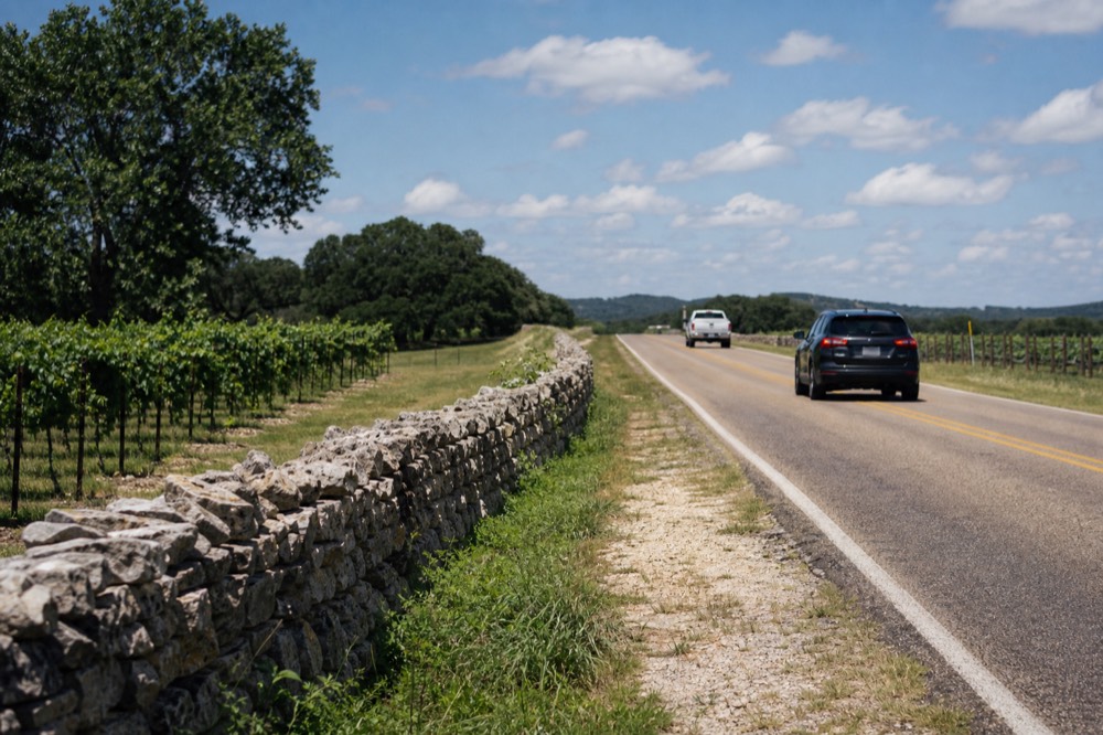 Two-lane Texas highway running past vineyard rows and a limestone fence line under a bright midday sky