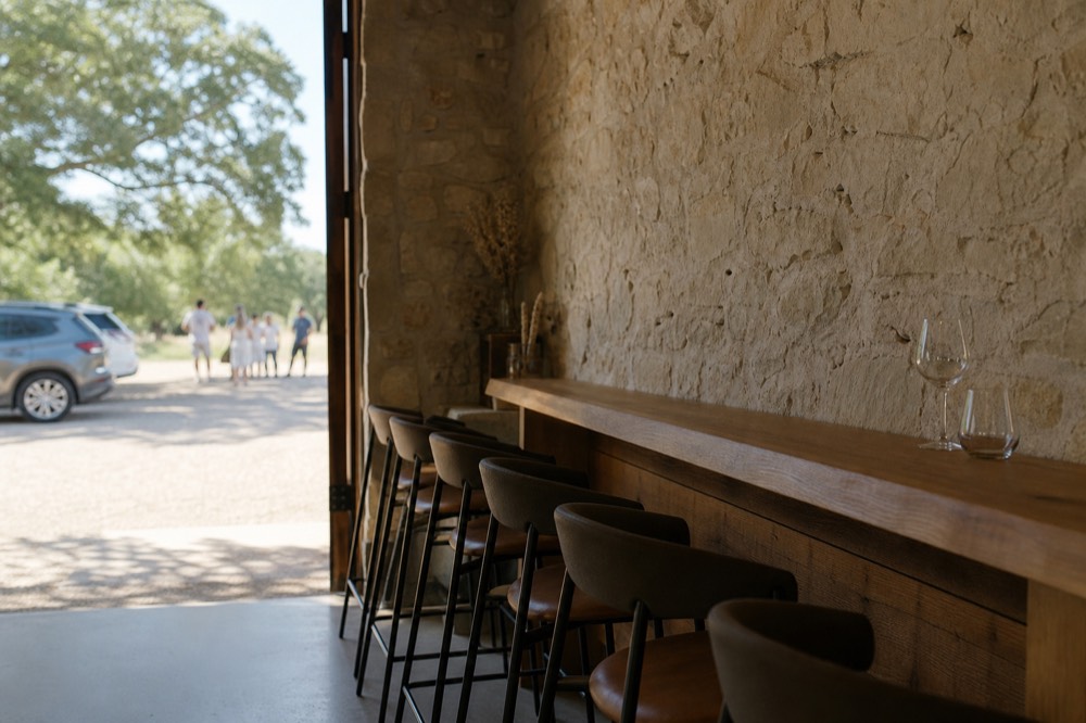 A small tasting room interior with a limestone wall, six bar stools and a wooden counter set with two glasses, empty and softly lit