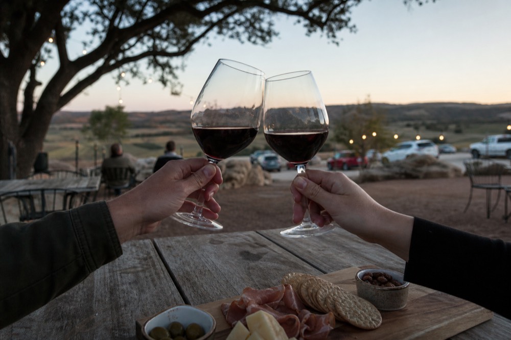 A couple's hands clinking two glasses of red wine over a rustic table at sunset, faces out of frame